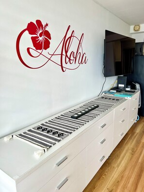 A room with a luggage carousel, a wall-mounted TV, and a wall decal with the word 'Aloha'. - Cozy studio with great view in fabulous Waikiki (Honolulu)