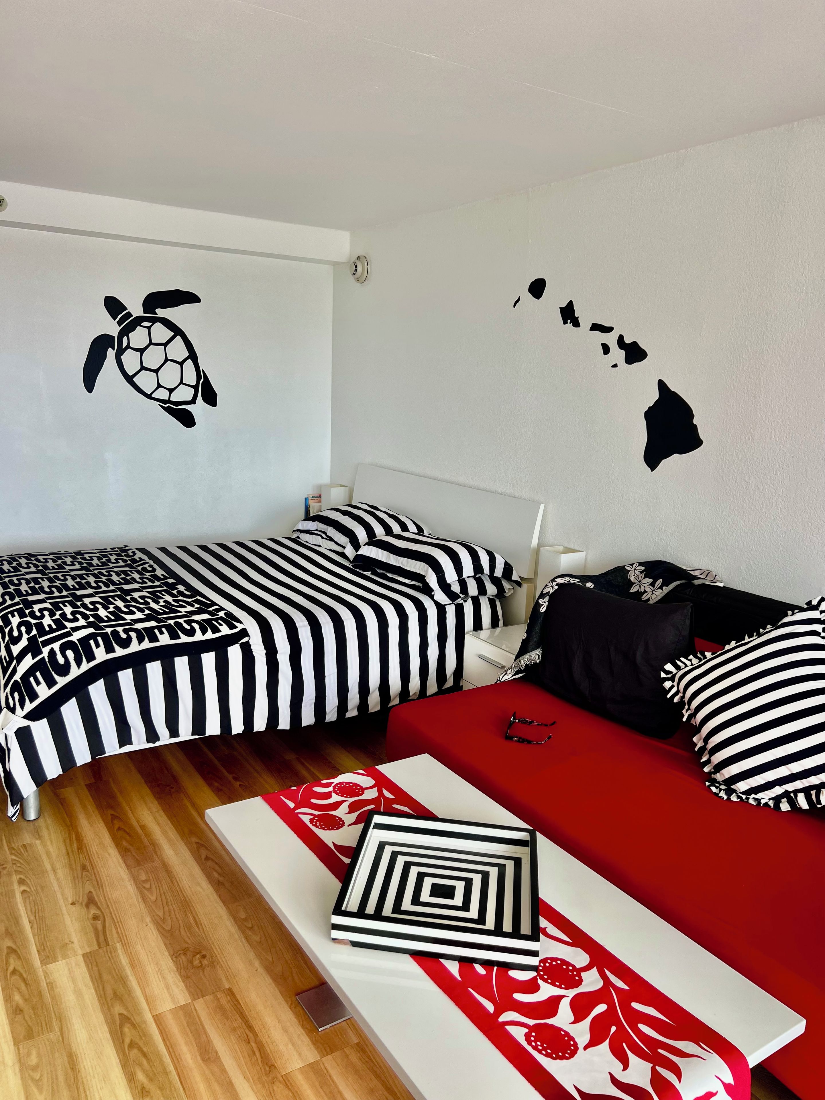 A room with two beds, one with striped bedding and the other with solid red bedding. There is a wall decal of a turtle and a map of Hawaii.