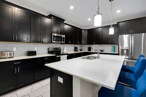 A modern kitchen with dark cabinetry, stainless steel appliances, a white countertop, and a blue chair. - Luxury Villa With Pool And Arcades (Four Corners)