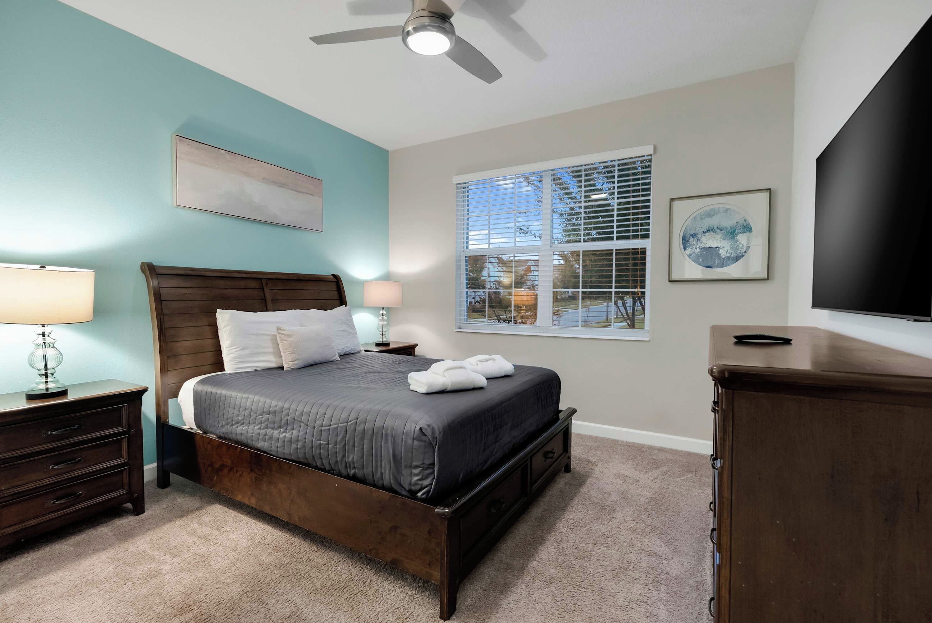 A bedroom with a bed, bedside tables, a ceiling fan, a window with blinds, and a flat-screen TV.