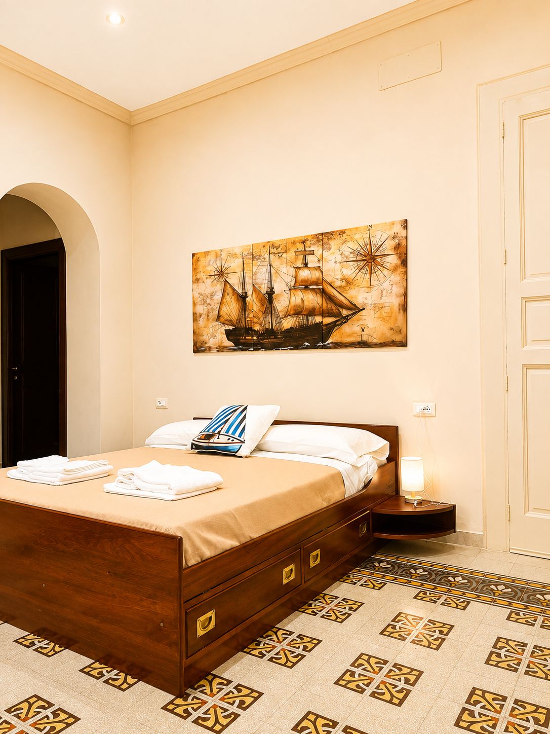 Quarto de hotel com cama, mesa de cabeceira, luminária e uma pintura grande na parede de um navio à vela.