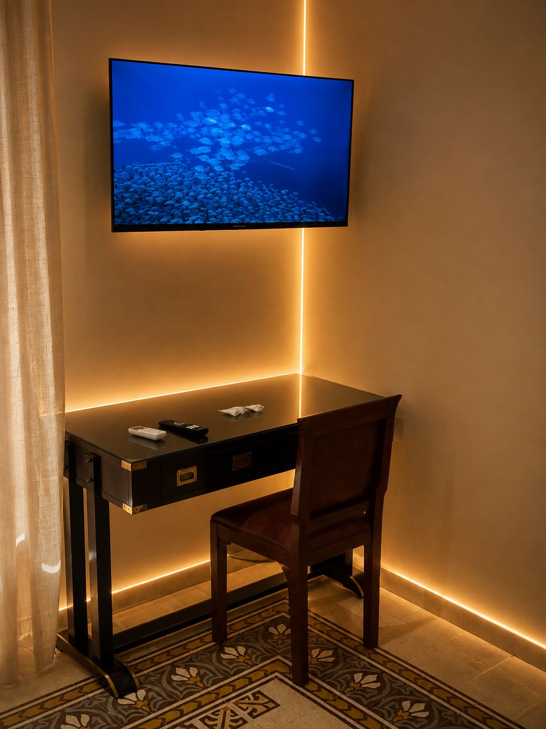 A hotel room with a flat-screen TV mounted on the wall, a wooden desk with a chair, and a patterned floor.