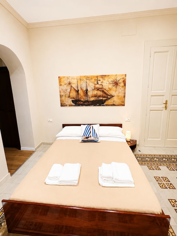 A hotel room with a large bed, a painting of a sailing ship, a nightstand with a lamp, and a door.
