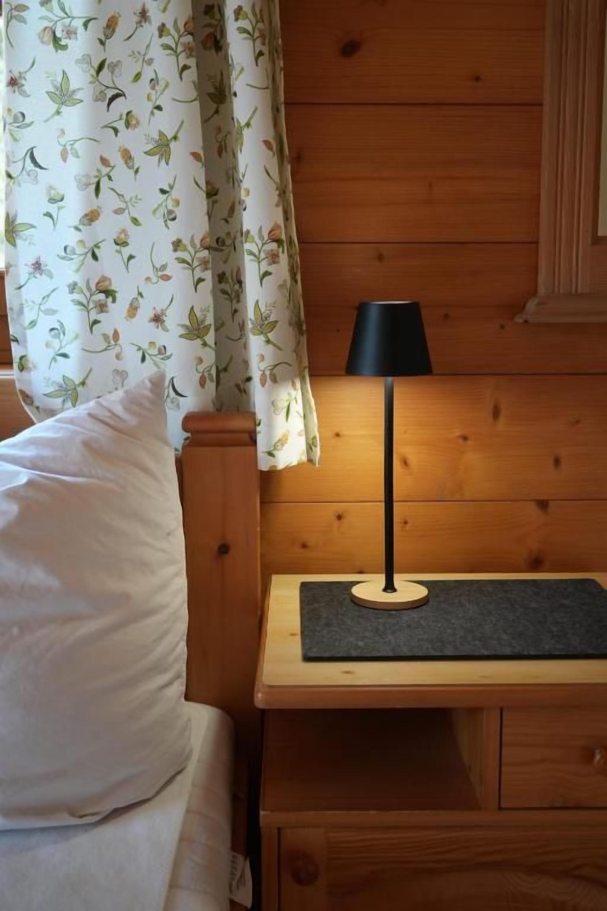 Una cama de madera con un almohadón blanco, una mesita de noche con una lámpara y una cortina estampada con motivos florales.