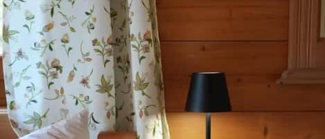 Una cama de madera con un almohadón blanco, una mesita de noche con una lámpara y una cortina estampada con motivos florales.