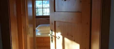 Un corridoio interno in legno che conduce a una stanza con una finestra, una porta con il chiavistello e un piccolo tappeto sul pavimento.