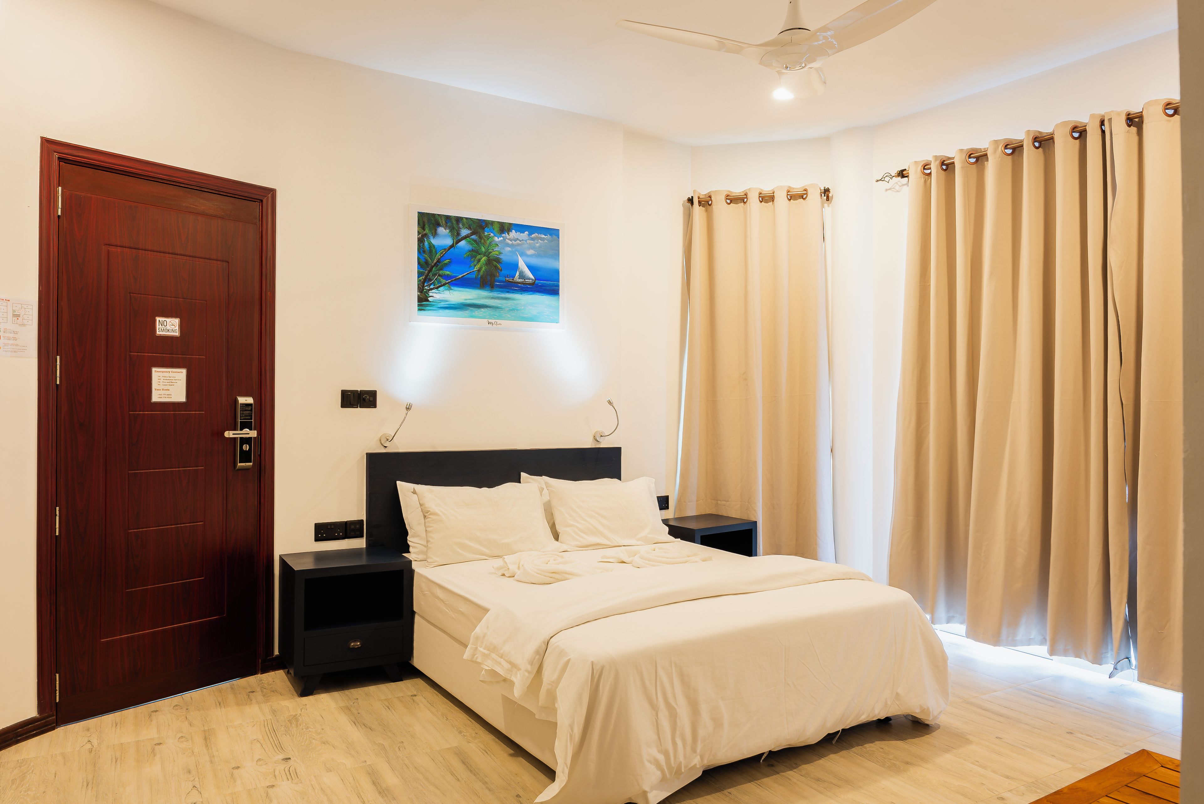 Una habitación de hotel con cama, mesita de noche, un cuadro en la pared, una puerta y cortinas.