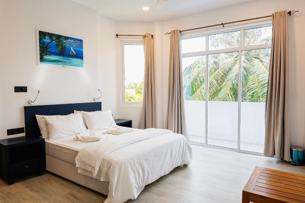 Habitación de hotel con cama, mesita de noche, ventana con cortinas y una pintura en la pared, con vista a palmeras.