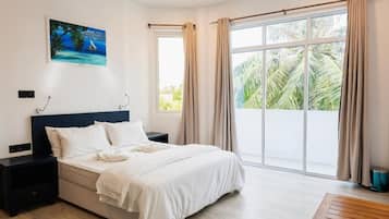 Habitación de hotel con cama, mesita de noche, ventana con cortinas y una pintura en la pared, con vista a palmeras.