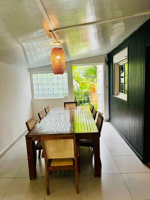 A dining area with a wooden table and chairs, a hanging light fixture, and a view of greenery outside. - C o s y 23, belle maison T4 de 108 m² avec jardin, supérettes et marchés locaux (Petite-Île)