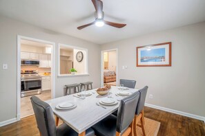 Un coin repas avec une table dressée pour quatre personnes, un ventilateur de plafond et une cuisine en arrière-plan.