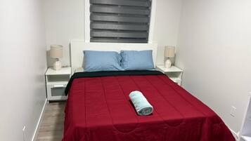A bedroom with a bed, two bedside tables, a window with blinds, and a rolled towel on the bed.