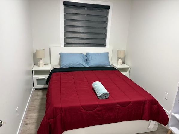 A bedroom with a bed, two bedside tables, a window with blinds, and a rolled towel on the bed.