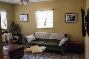 Un salon avec un canapé vert, un fauteuil en cuir marron, une table basse en bois et deux fenêtres ornées de rideaux légers.