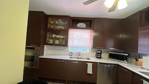 A kitchen with wooden cabinets, a stainless steel dishwasher, a microwave, and a window with blinds. - Iconic Wynwood Artsy Bungalow - Walk to Everywhere (Miami)