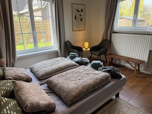 Et soveværelse med seng, puder, en stol, et lille bord og et vindue med gardiner.