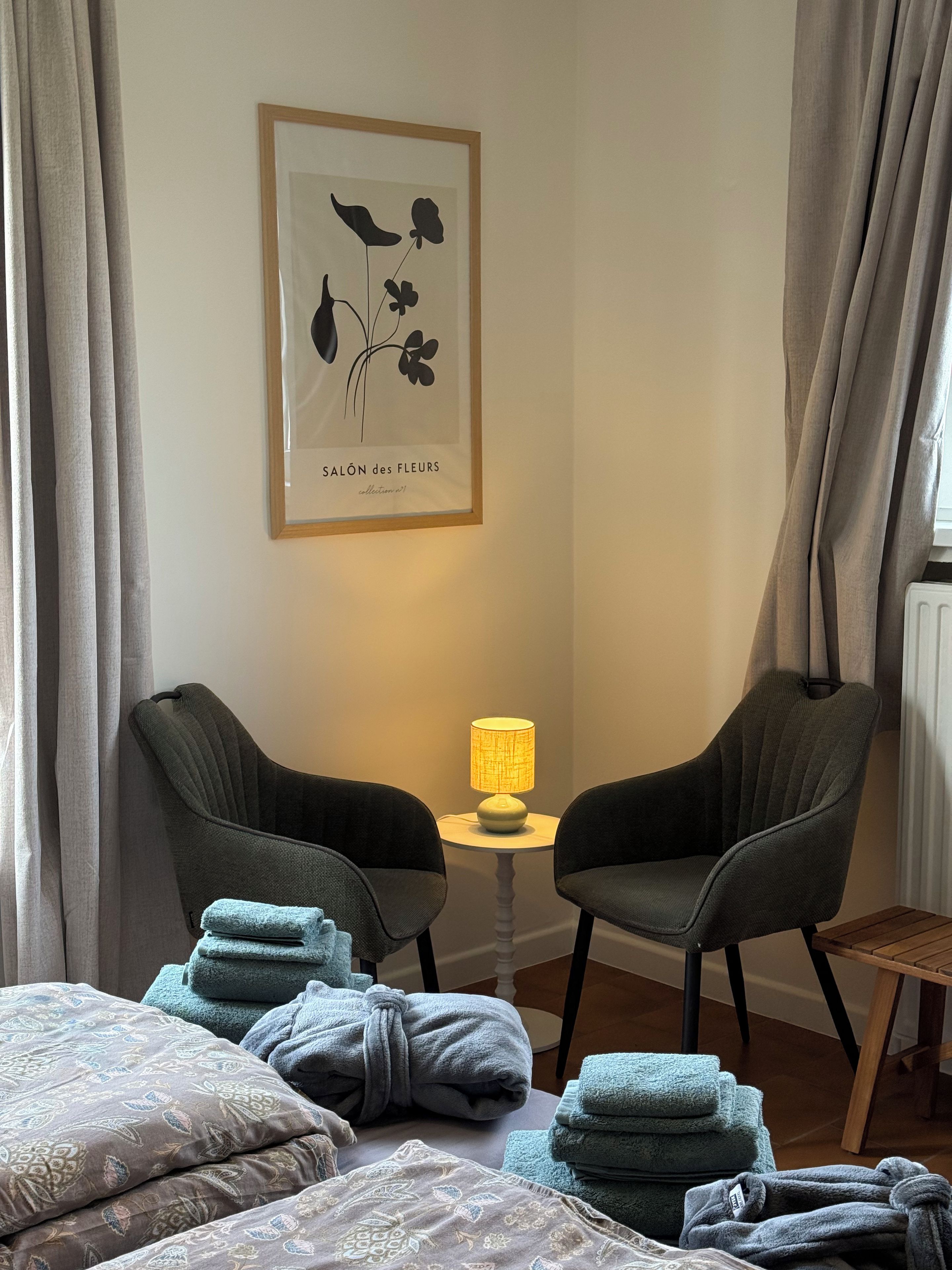 Una stanza con due sedie, un tavolino con una lampada e un letto con asciugamani e cuscini.