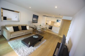 Ein modernes Wohnzimmer mit einer Couch, einem Couchtisch und einem Fernseher. Im Hintergrund ist eine Küche mit Essbereich zu sehen.