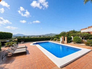 Ein Swimmingpool mit gefliestem Deck und Liegestühlen, umgeben von Grün und Blick auf ferne Hügel.