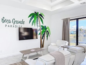 A modern living room with a flat-screen TV, white leather sofa, a coffee table, and a large window with a view of a body of water. - Boca Grande Retreat Sleeps 10 (Port Charlotte)