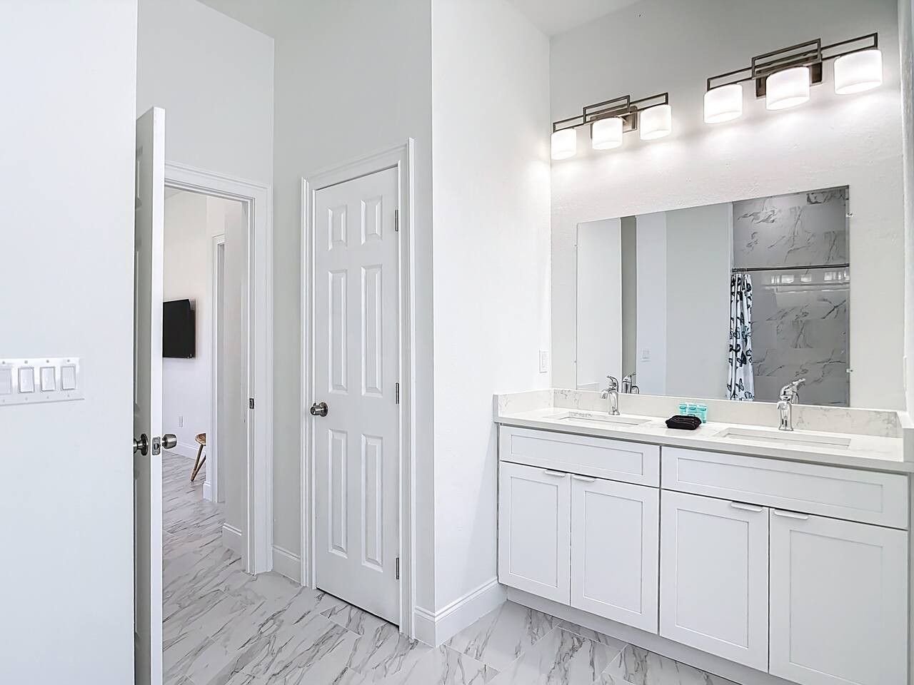 Une salle de bain avec un meuble-lavabo à deux vasques, un grand miroir et une porte donnant sur une autre pièce.