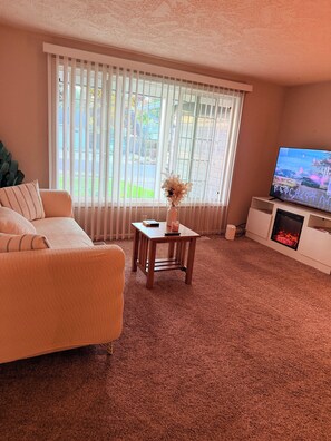 A living room with a beige sofa, a small wooden table with a vase, a flat-screen TV, and a fireplace. - Gorgeous 3-bedroom house with AC in charming Eugene (Eugene)