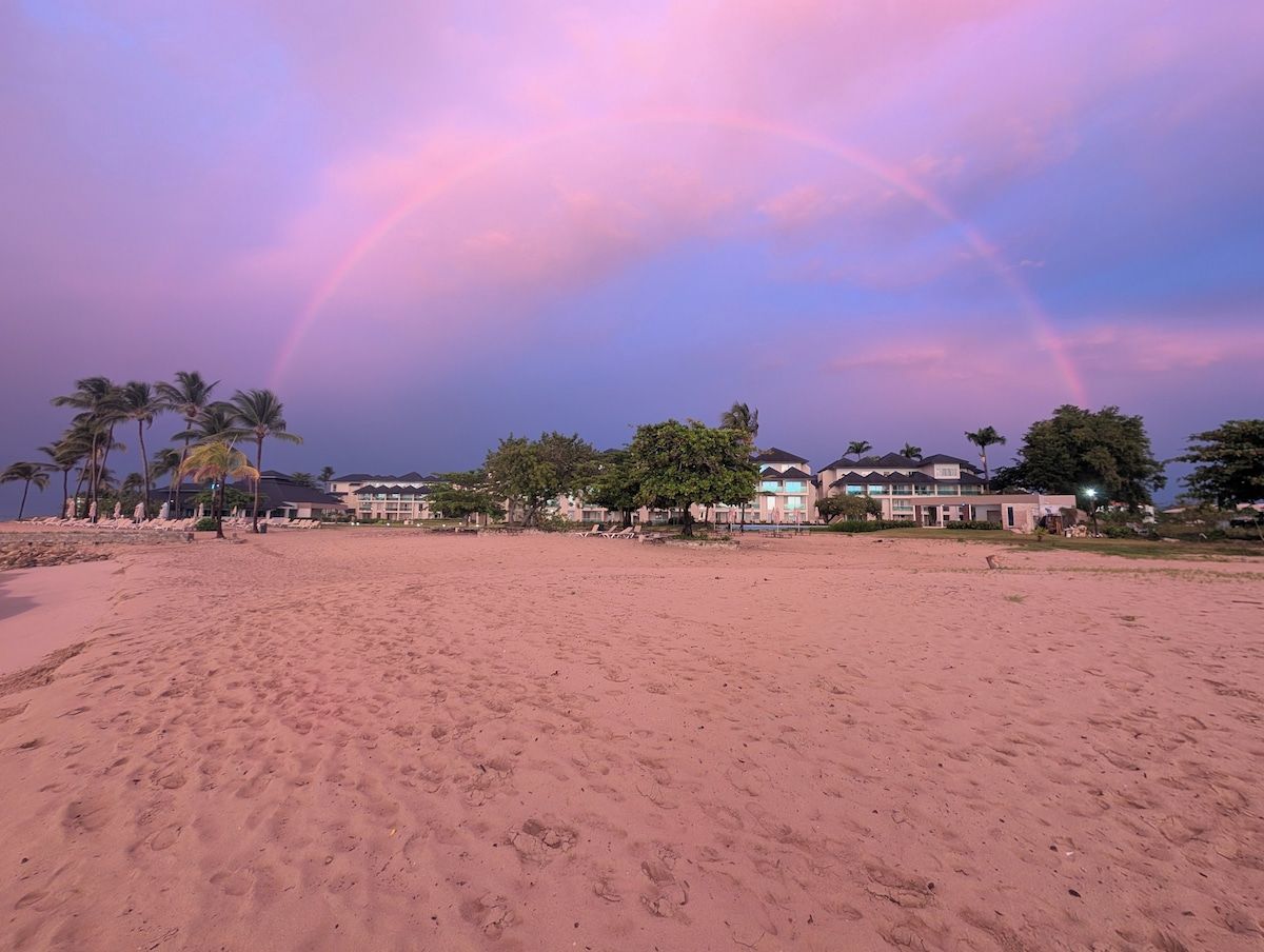 Una playa con cielo despejado y un arcoíris. Se ven palmeras y edificios al fondo.