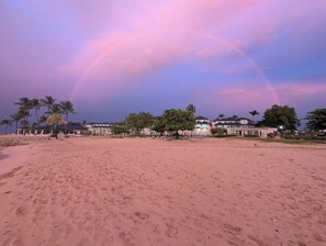 Une plage avec un ciel dégagé et un arc-en-ciel. On aperçoit des palmiers et des bâtiments au loin.