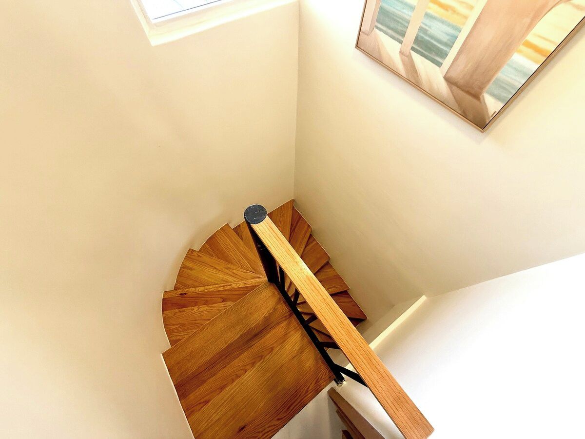 A wooden spiral staircase with a handrail, a window above, and a painting on the wall.