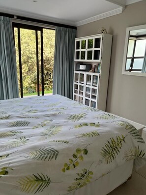 A bedroom with a large bed, a window with curtains, a white cabinet, and a view of greenery outside.