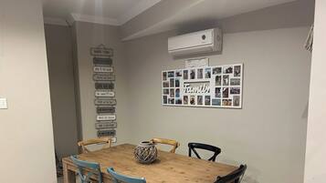 Un coin repas avec une table en bois et quatre chaises, un appareil de climatisation au-dessus, et un mur orné d’un collage de photos de famille.