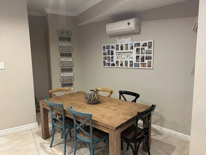 A dining area with a wooden table and four chairs, an air conditioning unit above, and a wall with a family photo collage.