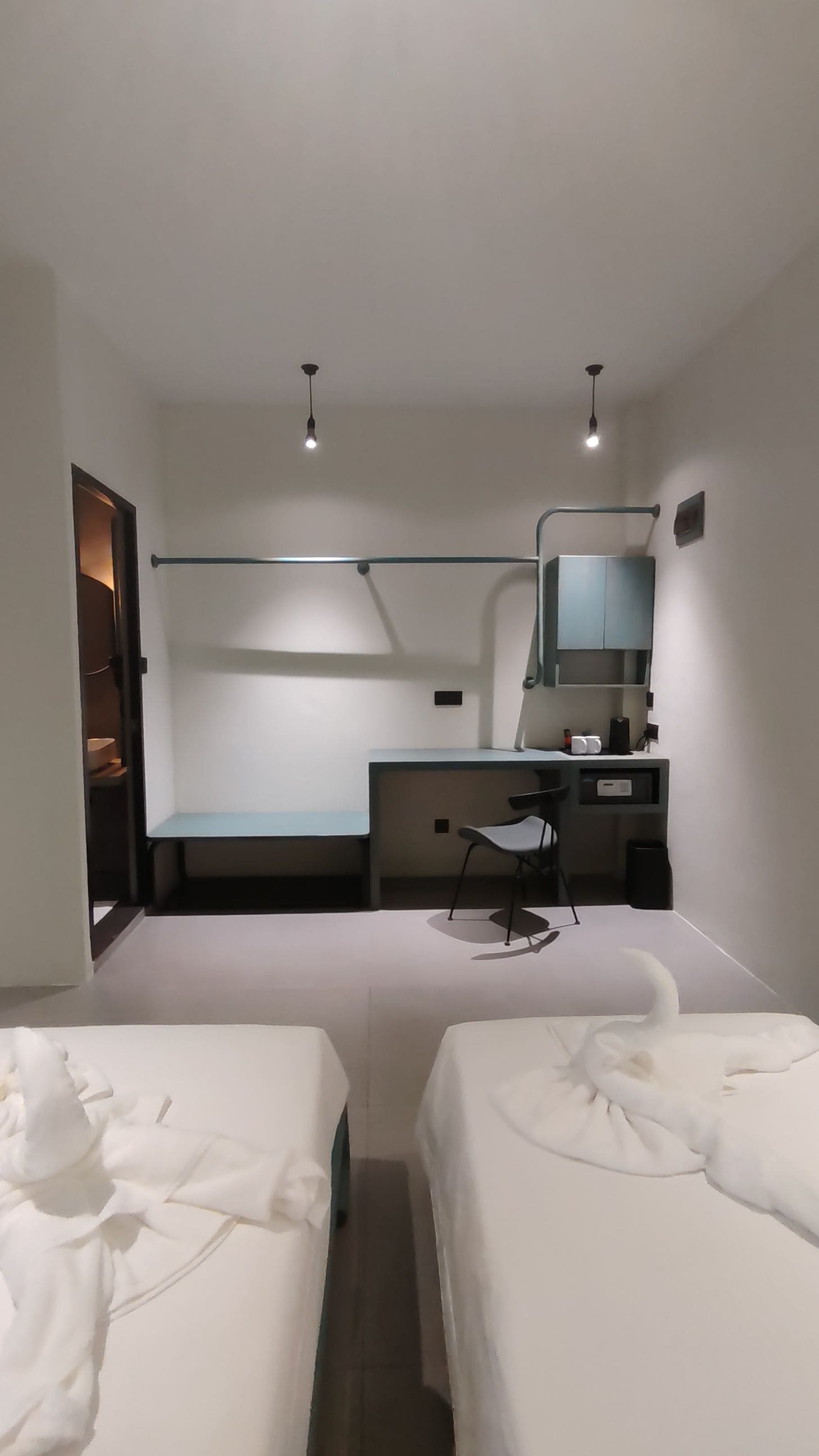Habitación de hotel con dos camas, un escritorio con silla, una mesita, un espejo y una lámpara colgante.