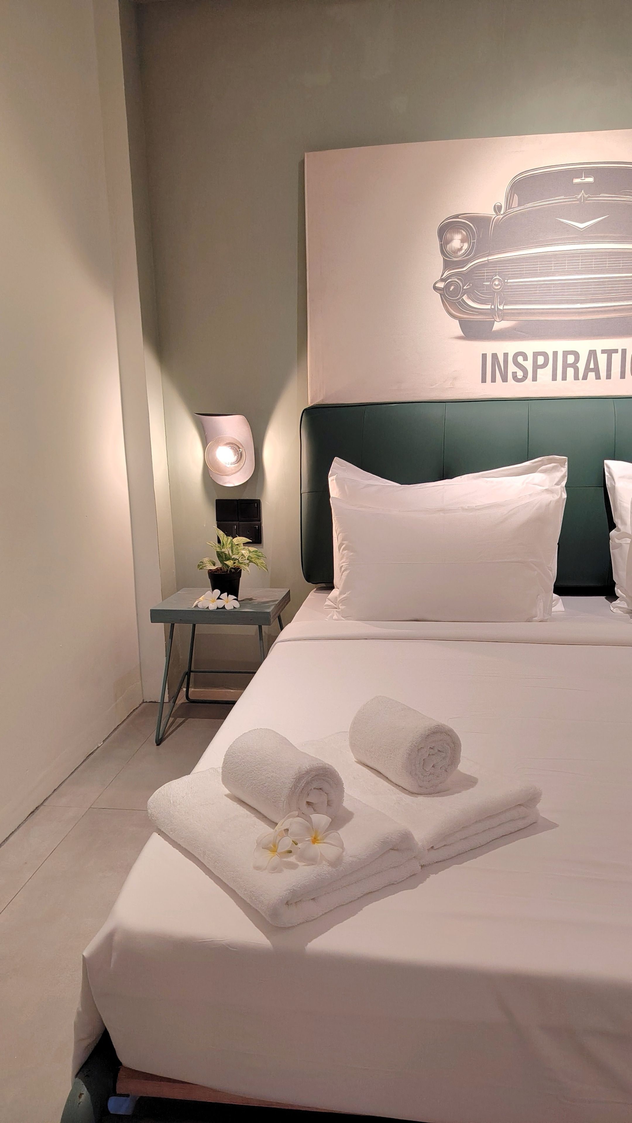 A hotel room with a bed, pillows, a nightstand with a lamp, a wall art piece, and rolled towels.