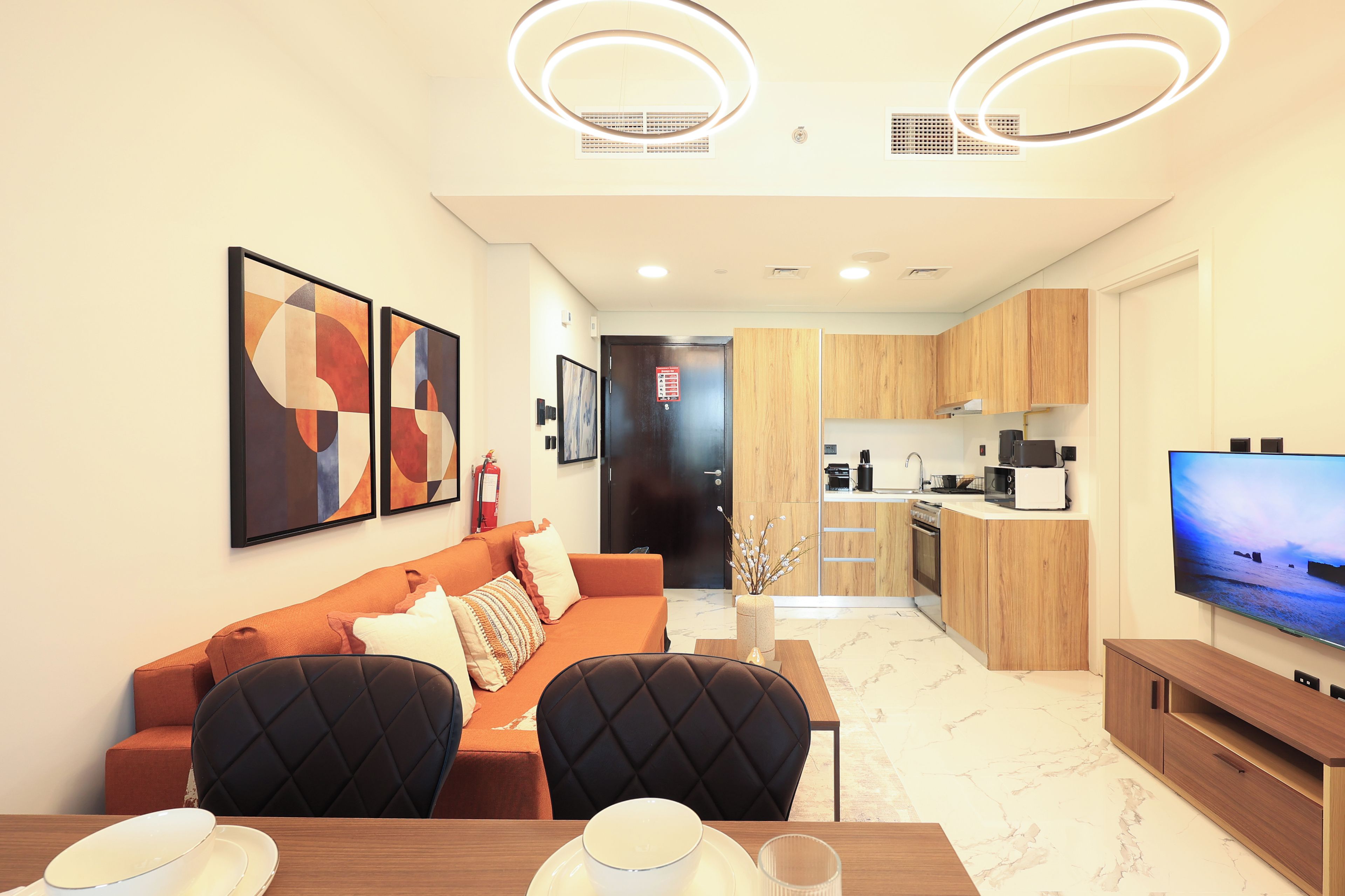 A modern living room with a sofa, chairs, a dining table, and a kitchen area with wooden cabinets and a flat-screen TV.