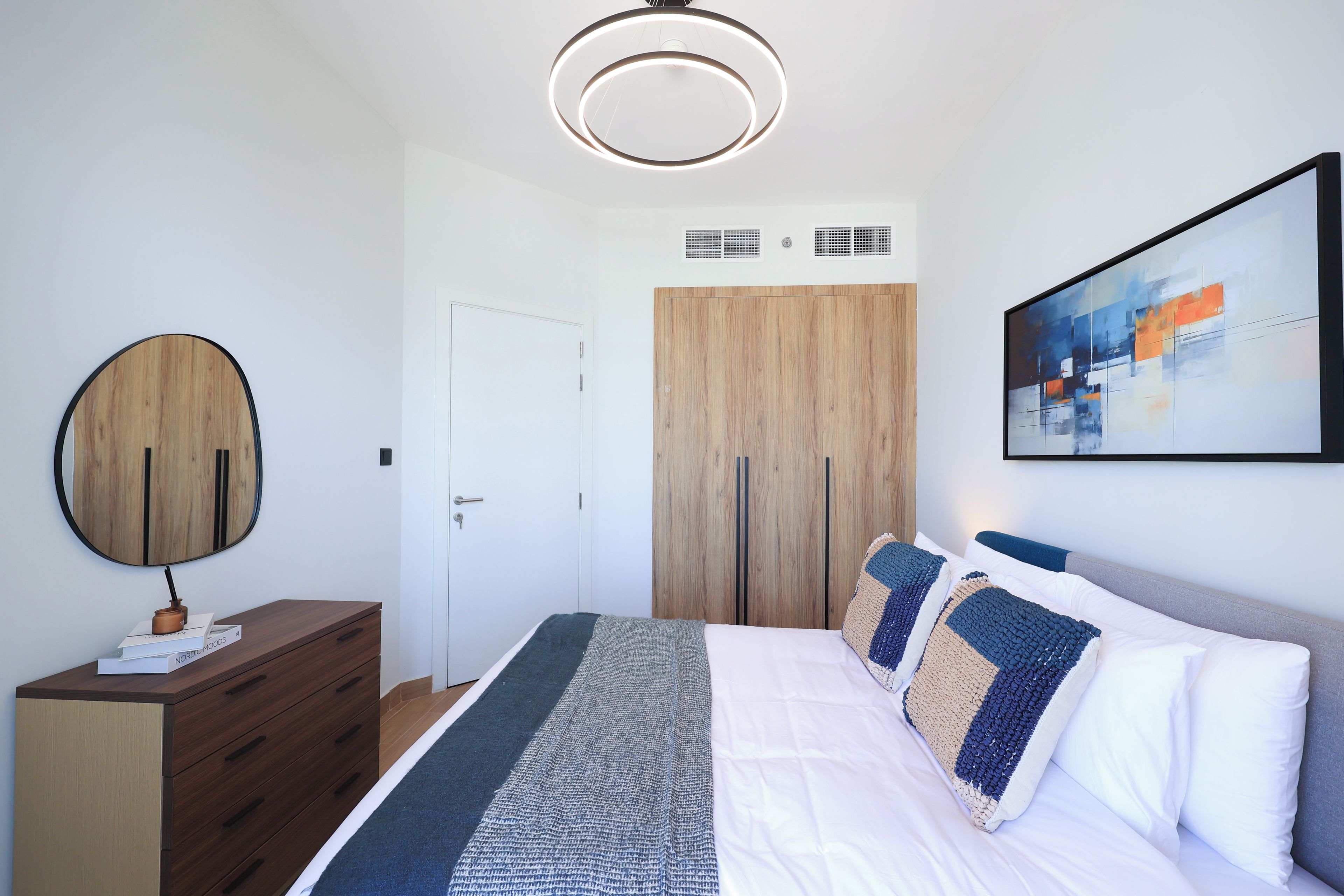 Un dormitorio moderno con cama, cómoda, espejo, un cuadro y una lámpara en el techo.