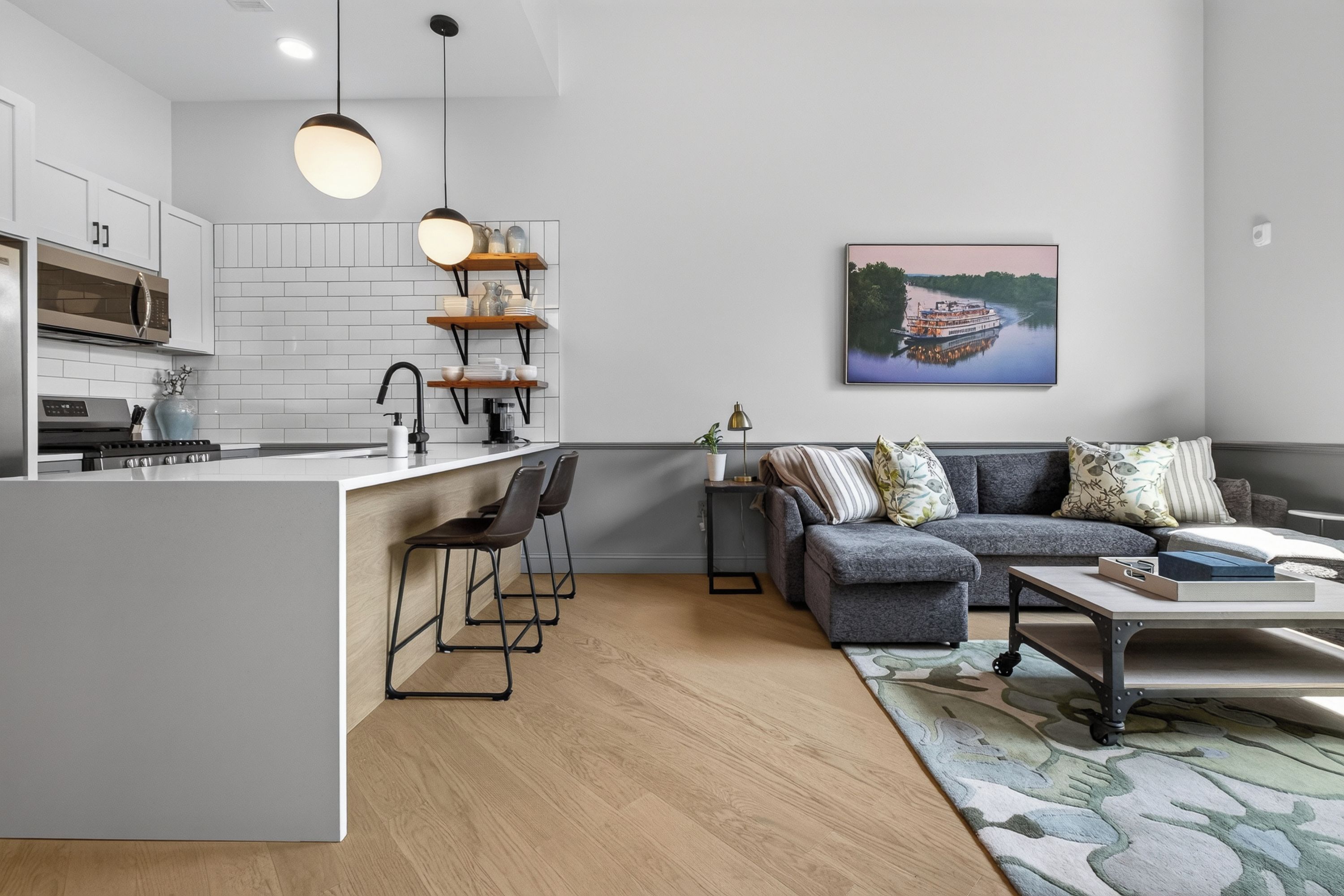 Eine moderne Küche mit Kochinsel, ein Essbereich mit grauem Sofa und Couchtisch sowie ein Wohnzimmer mit einem großen Gemälde einer Flusslandschaft.