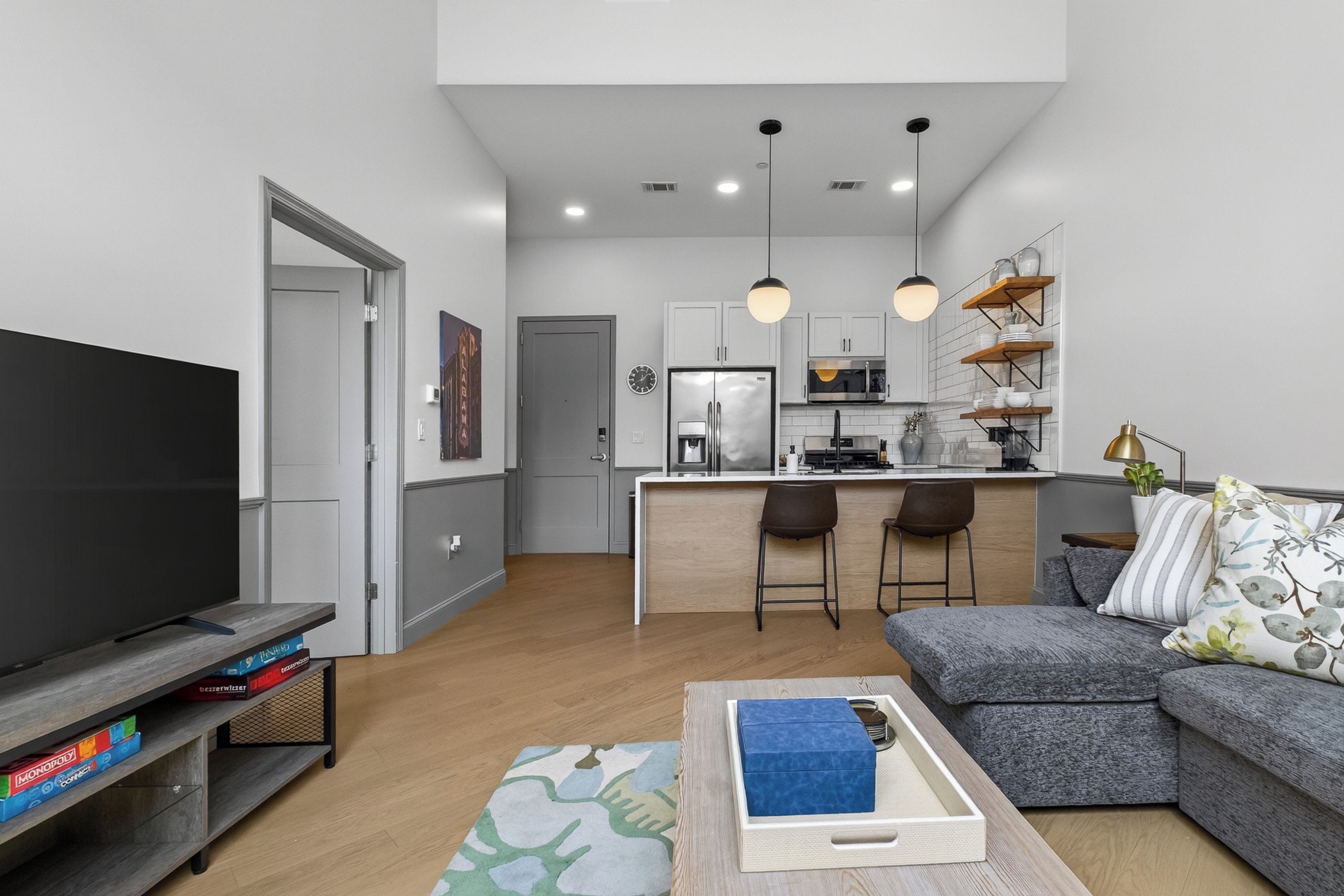 A modern living room with a flat-screen TV, a sofa, and a coffee table. There is a kitchen area with a refrigerator, microwave, and bar stools.