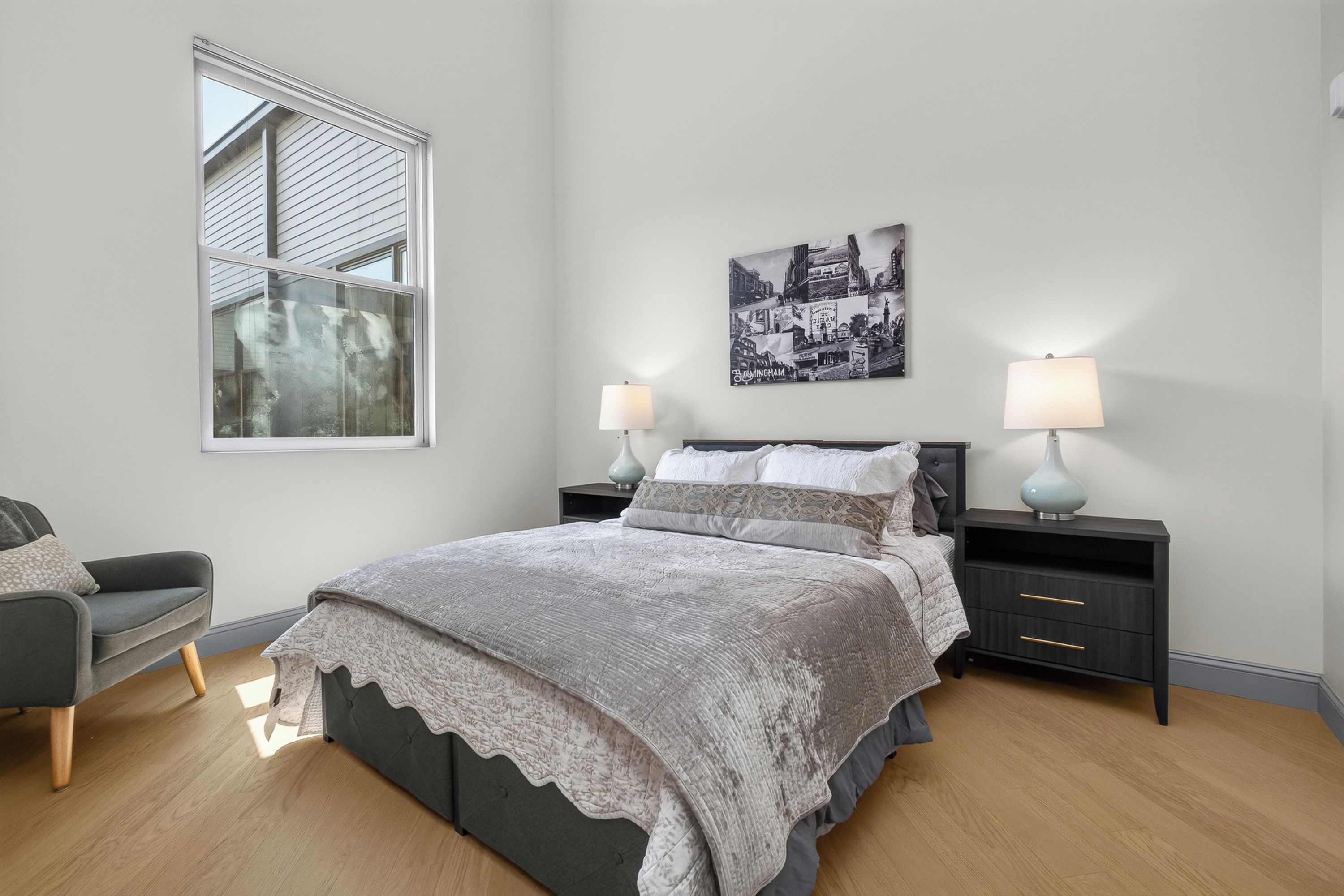 Une chambre à coucher comprenant un lit, deux tables de chevet, une chaise, une fenêtre et un tableau accroché au mur.