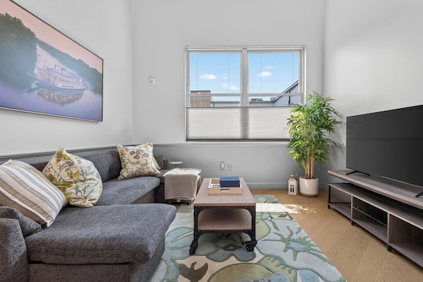 A living room with a grey sofa, a coffee table, a flat-screen TV, a potted plant, and a painting of a boat on the wall.