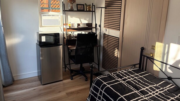 Ein kleines Schlafzimmer mit Bett, Schreibtisch, Stuhl, kleinem Kühlschrank und einem Fenster mit Jalousien.