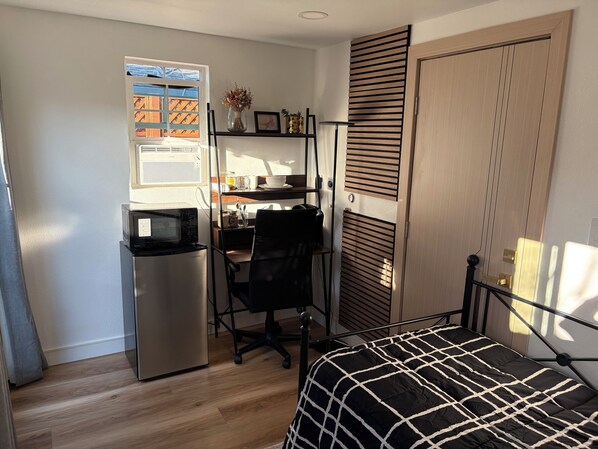 A compact bedroom with a bed, a desk, a chair, a small refrigerator, and a window with blinds.