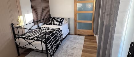 Quarto com cama, janela com persianas, porta e piso de madeira.