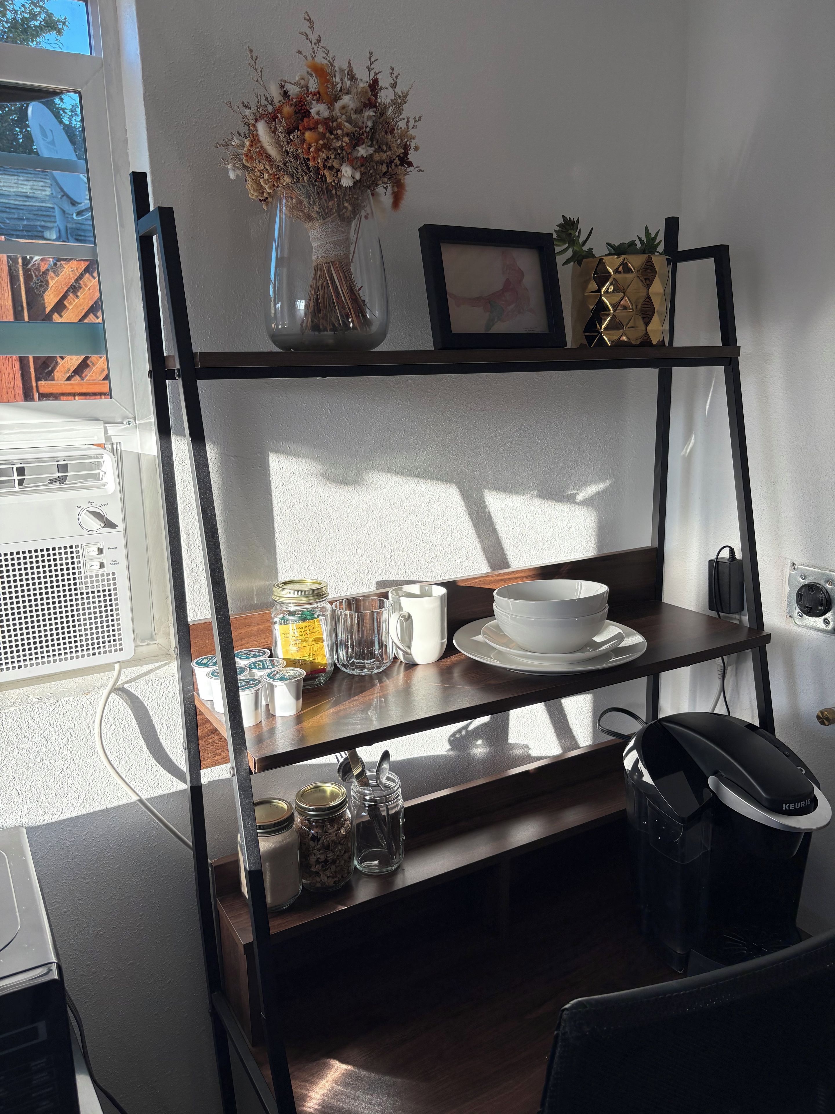 Una zona de cuina amb una estant que conté un ram de flors seques, un quadre amb marc i una petita planta en test. Hi ha pots, una cafetera i un bullidor a la tauleta.