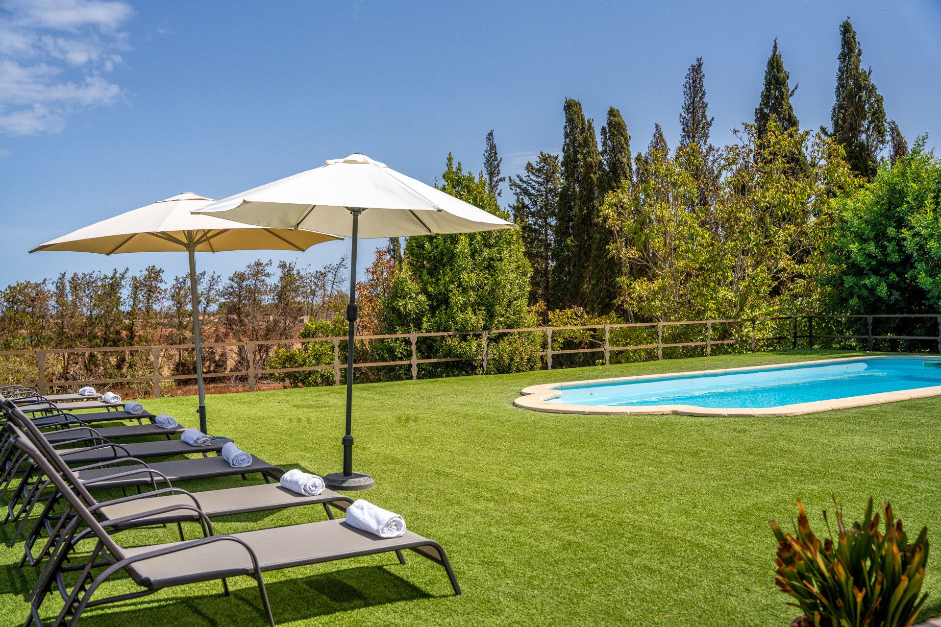 A poolside area with lounge chairs and umbrellas, surrounded by a wooden fence and trees.
