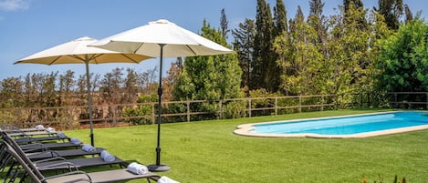 Zona junto a la piscina con sillones de descanso y sombrillas, rodeada por una valla de madera y árboles.