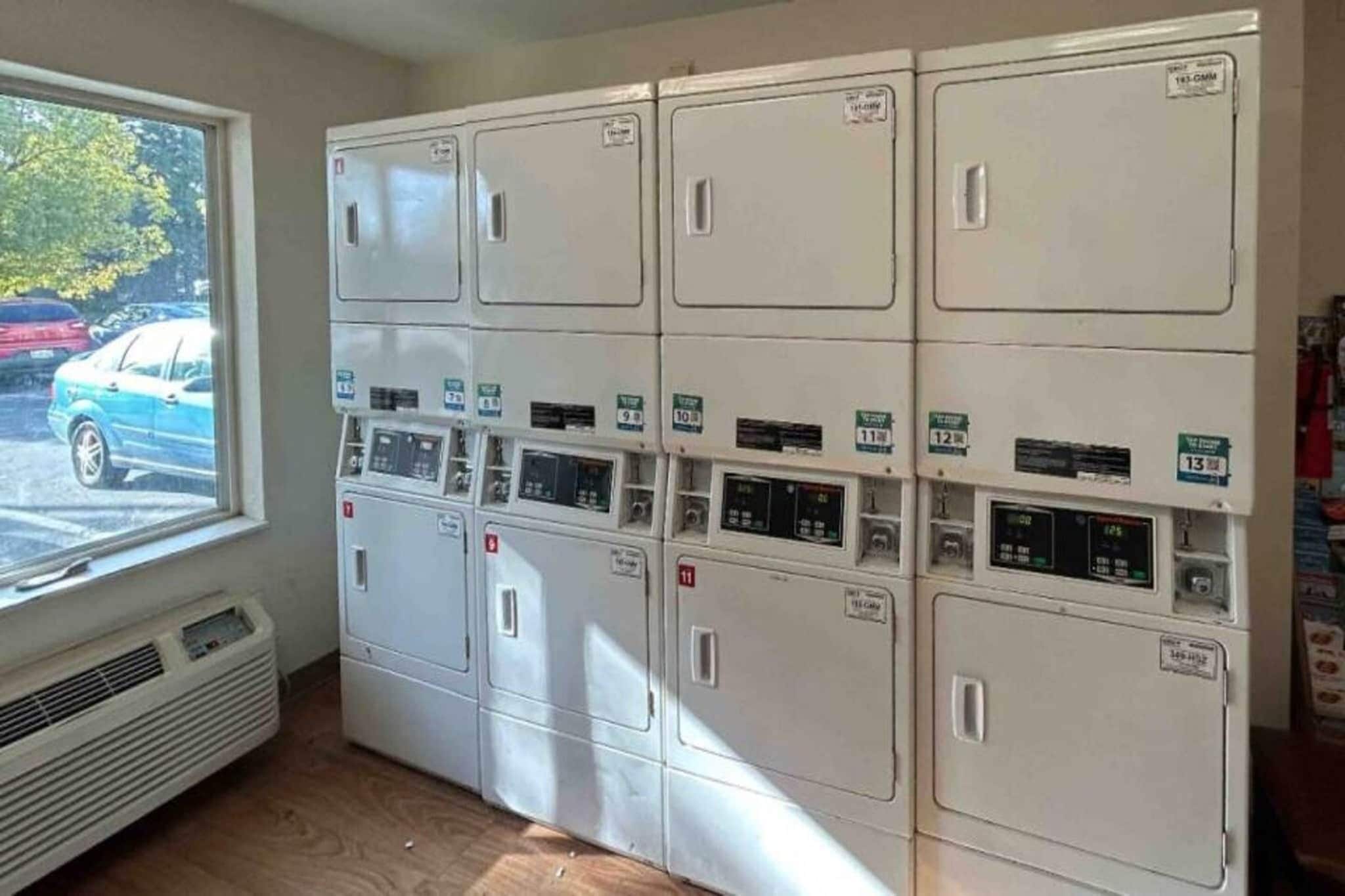 A laundry room with multiple washing machines and dryers, a window with a view of parked cars, and a vent on the wall.