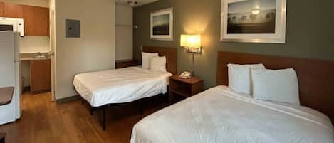 A hotel room with two beds, a desk, a chair, a small kitchenette, and a painting on the wall.