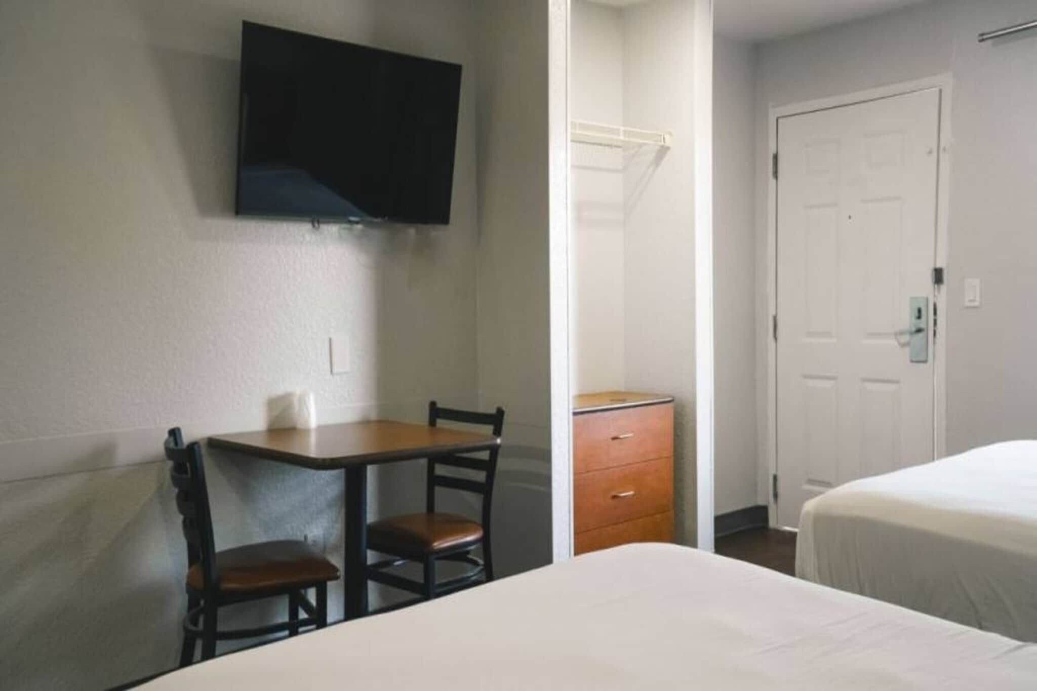 A hotel room with a bed, a desk with two chairs, a television mounted on the wall, and a door.