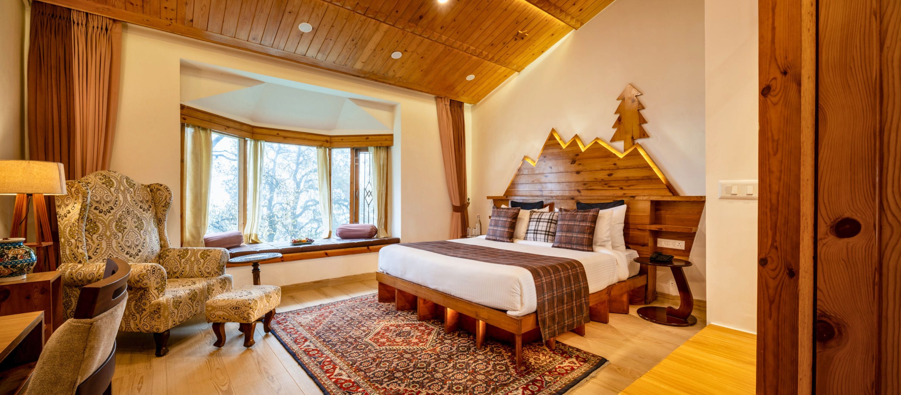 Ein Schlafzimmer mit Holzboden, einem Bett mit Holz Kopfteil, einem Fenster mit Vorhängen, einem gemusterten Sessel und einem kleinen Tisch mit Lampe.